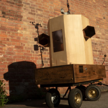 looking up at the Future Machine, brass wheels, oak base with brass intructions and button on the side, copper trumpets on each side of an ash octagon with a copper panel in the front and a pole at the back, shadow of the weather station at the top against a red brick wall