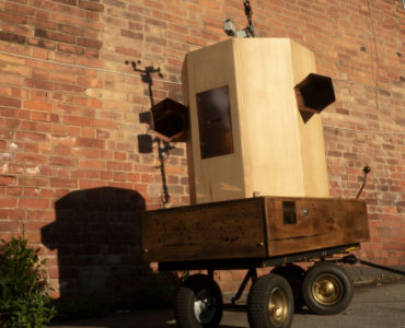 looking up at the Future Machine, brass wheels, oak base with brass intructions and button on the side, copper trumpets on each side of an ash octagon with a copper panel in the front and a pole at the back, shadow of the weather station at the top against a red brick wall