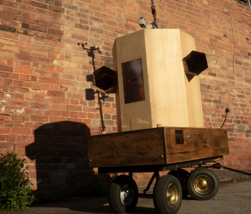 looking up at the Future Machine, brass wheels, oak base with brass intructions and button on the side, copper trumpets on each side of an ash octagon with a copper panel in the front and a pole at the back, shadow of the weather station at the top against a red brick wall