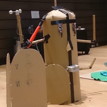 Future Machine prototype - a large cardboard head with sensors for eyes and a rainfall sensor for a mouth with 4 arms sticking up in the air and a round door.