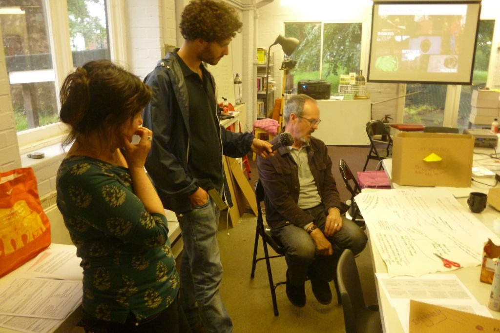 Rachel and a participant in workshop being recorded on a microphone by another participant in Furtherfield Commons, screen with images in the background