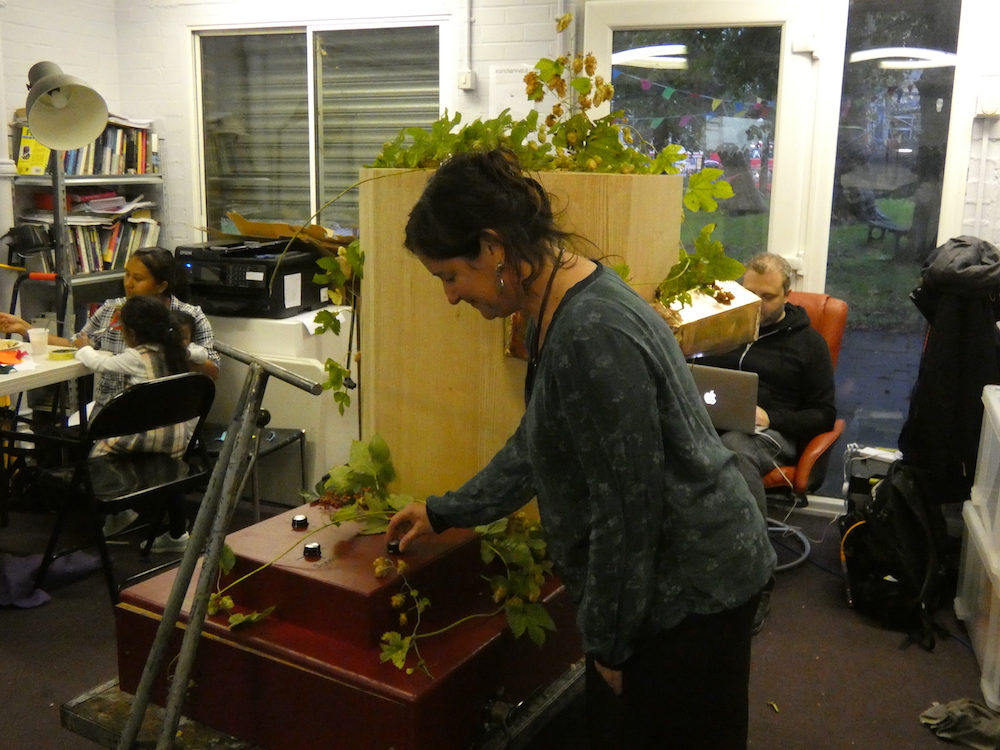 Rachel turning a dial on the Future Machine, decorated with hops in Furtherfield commons with people sitting behind