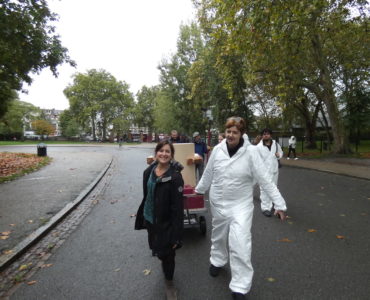 Rachel Jacobs and Ruth Catlow pulling the Future Machine uphill with the procession of people behind and Autumnal trees in Finsbury Park