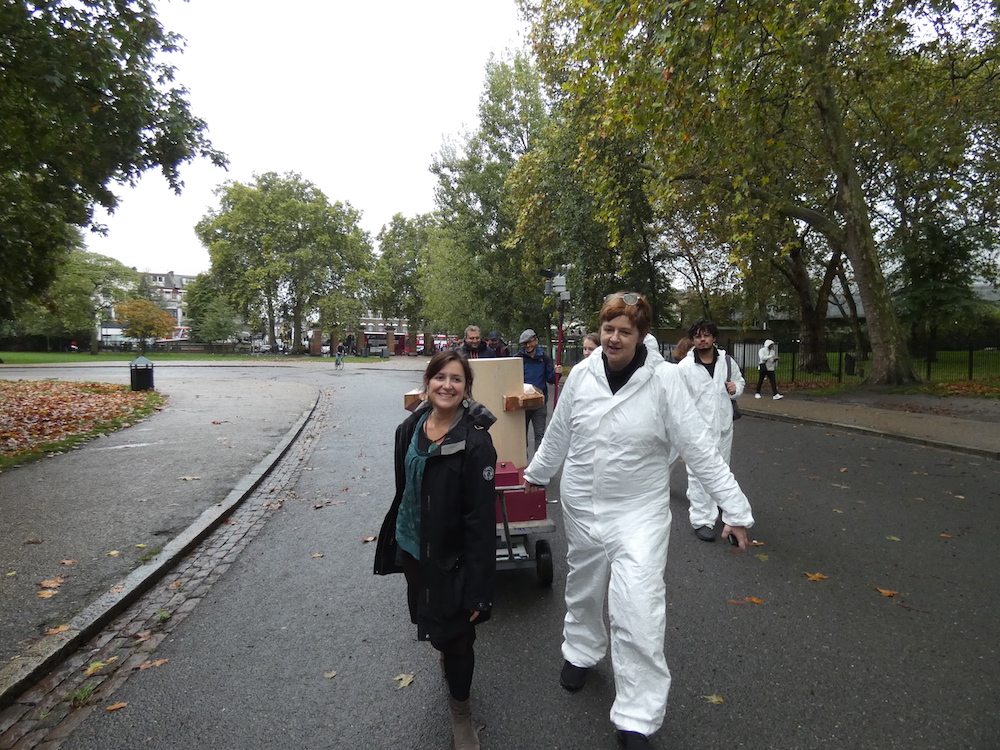 Rachel Jacobs and Ruth Catlow pulling the Future Machine uphill with the procession of people behind and Autumnal trees in Finsbury Park