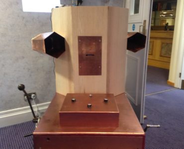 Future Machine with it's copper coloured trolley, box of dials, lever, handcrank and the Octagon with a copper rectangle in the front with a slot for the printed messages to come out and two copper trumpets on either side