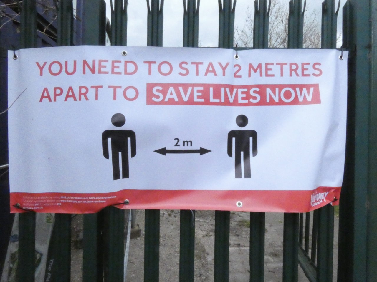 banner tied to metal fence saying You need to stay 2 metres apart to save lives now