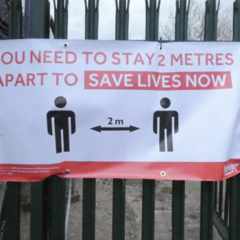 banner tied to metal fence saying You need to stay 2 metres apart to save lives now