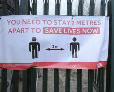 banner tied to metal fence saying You need to stay 2 metres apart to save lives now