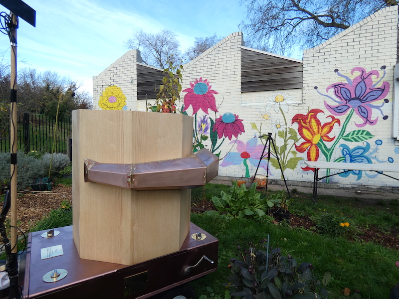 side of the Future Machine in the drumming school garden with the mural behind and the lever and weather pole, the drumming school garden and mural in the background