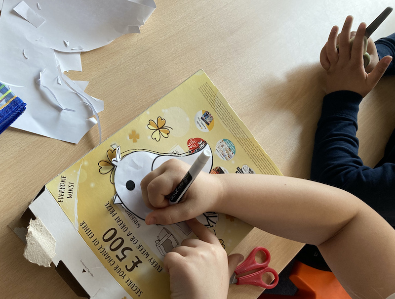 A child's hands drawing an outline of a bird onto a cereal packet, with scissors in the background and another childs hands holding scissors