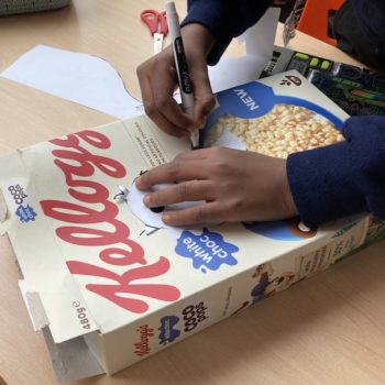 A child's hands drawing an outline of a bird onto a Kellogs Coco Pops cereal packet, with scissors in the background