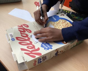 A child's hands drawing an outline of a bird onto a Kellogs Coco Pops cereal packet, with scissors in the background