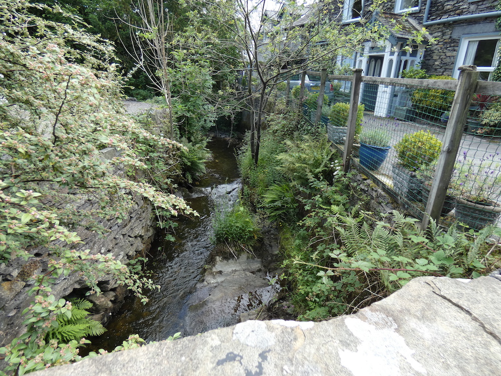 river along the side of a house with a metal and wooden fence, fern, slate, dry stone wall on the otherside and trees
