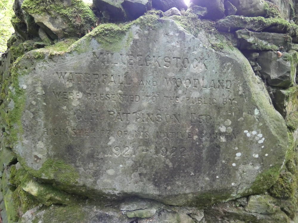 carved into a mossy slate rock with a dry stone wall behind - Millbeck Stock Waterfall and Woodland were presented to the public by D.H. Pattinson Esq High (unreadable) of his native county 1921 - 1922