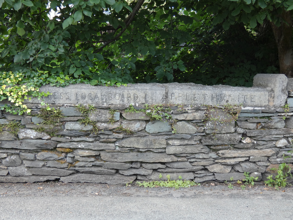 a road, a dry stone wall with engraved writing into the top of the wall (unreadable), trees