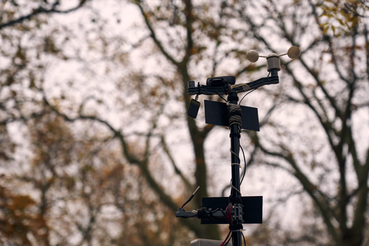 wind speed, temperature and moisture sensors with solar panels and a webcam on a pole with cables wrapped around and the Autumn trees blurred in the distance