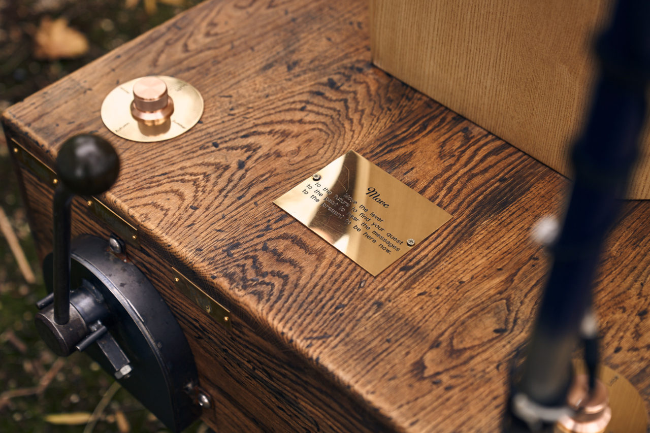 distressed oak base of the Future Machine with the bottom of the weather pole in the foreground, a brass sign that says Move and instructions blurred, two brass dials with brass discs with blurred writing on, a large metal lever, the bottom of the octagon and leaves on the floor below