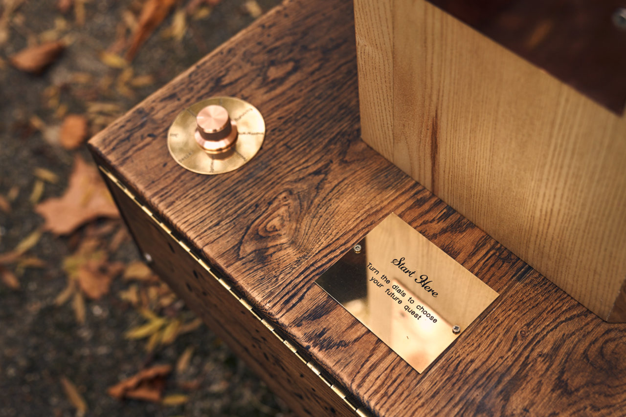 looking down at the front of Future Machine, a brass sign with Start Here - turn the dials to choose your future quest. Distressed oak base, the ash octagon and a brass dial