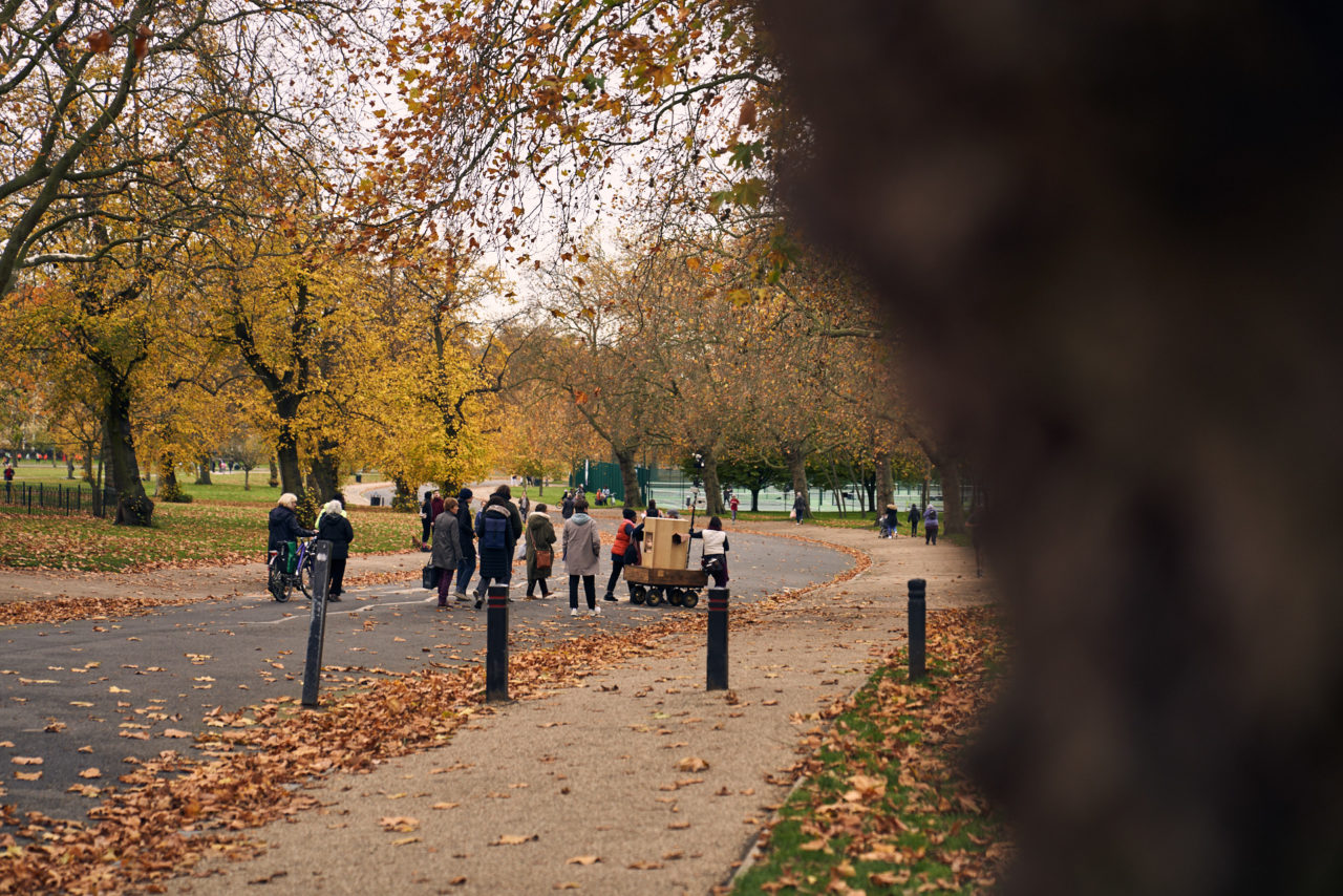 A tree trunk in the foreground and the Future Machine and procession of 9 people in the distance walking down hill surrounded by autumnal trees