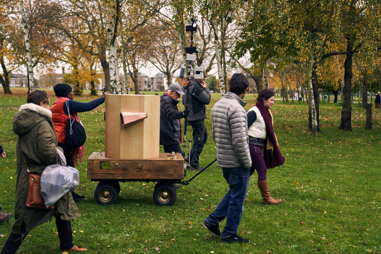 Esi pushing, Rachel pulling the Future Machine, man holding the weather pole, two people walking alongside, autumnal trees, grass and buildings in the distance on Seven Sisters Road