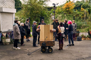 May DeGrace talking to the procession outside the drumming school with Future Machine in the foreground