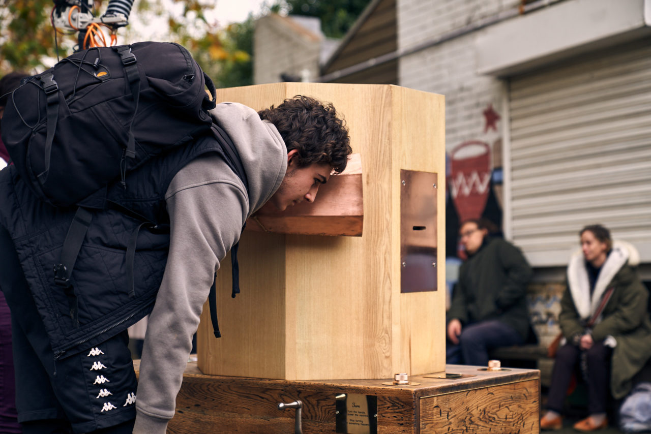 a young man listening to the copper trumpet of the Future Machine with people in the background sitting on a bench outside the drumming school