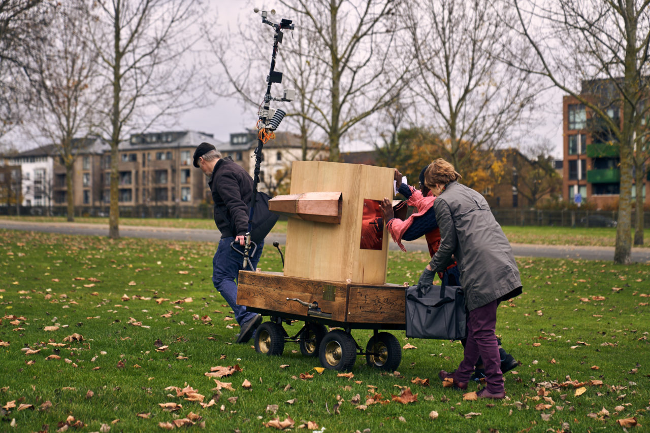 A man and two women pulling and pushing the Future Machine up a grassy hill with buildings on Seven Sisters Road in the background