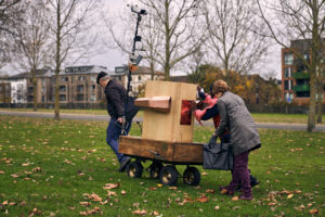 A man and two women pulling and pushing the Future Machine up a grassy hill with buildings on Seven Sisters Road in the background