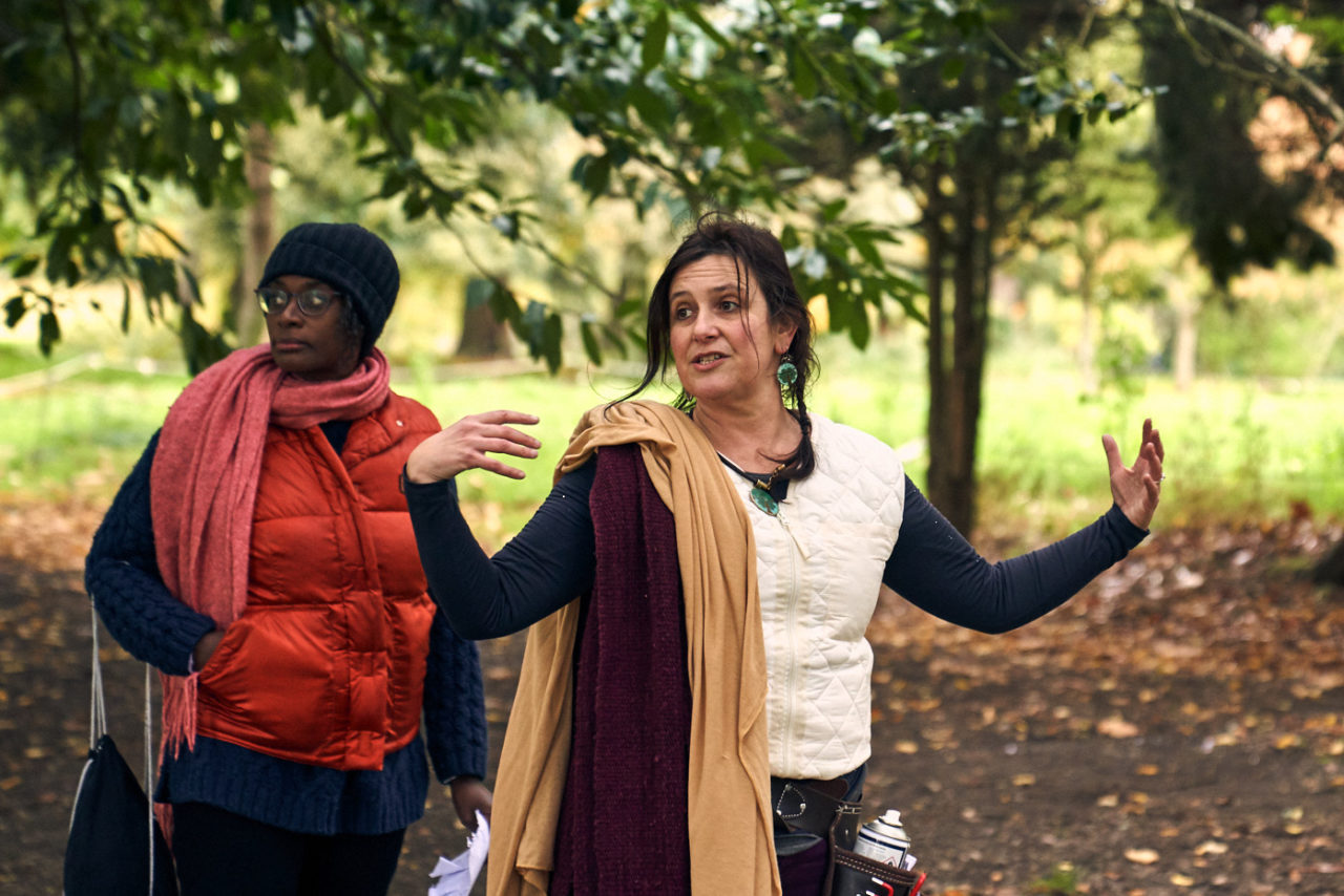 Rachel Jacobs talking with her arms in the air, with Esi Eshun behind and rodedendrum leaves in the background