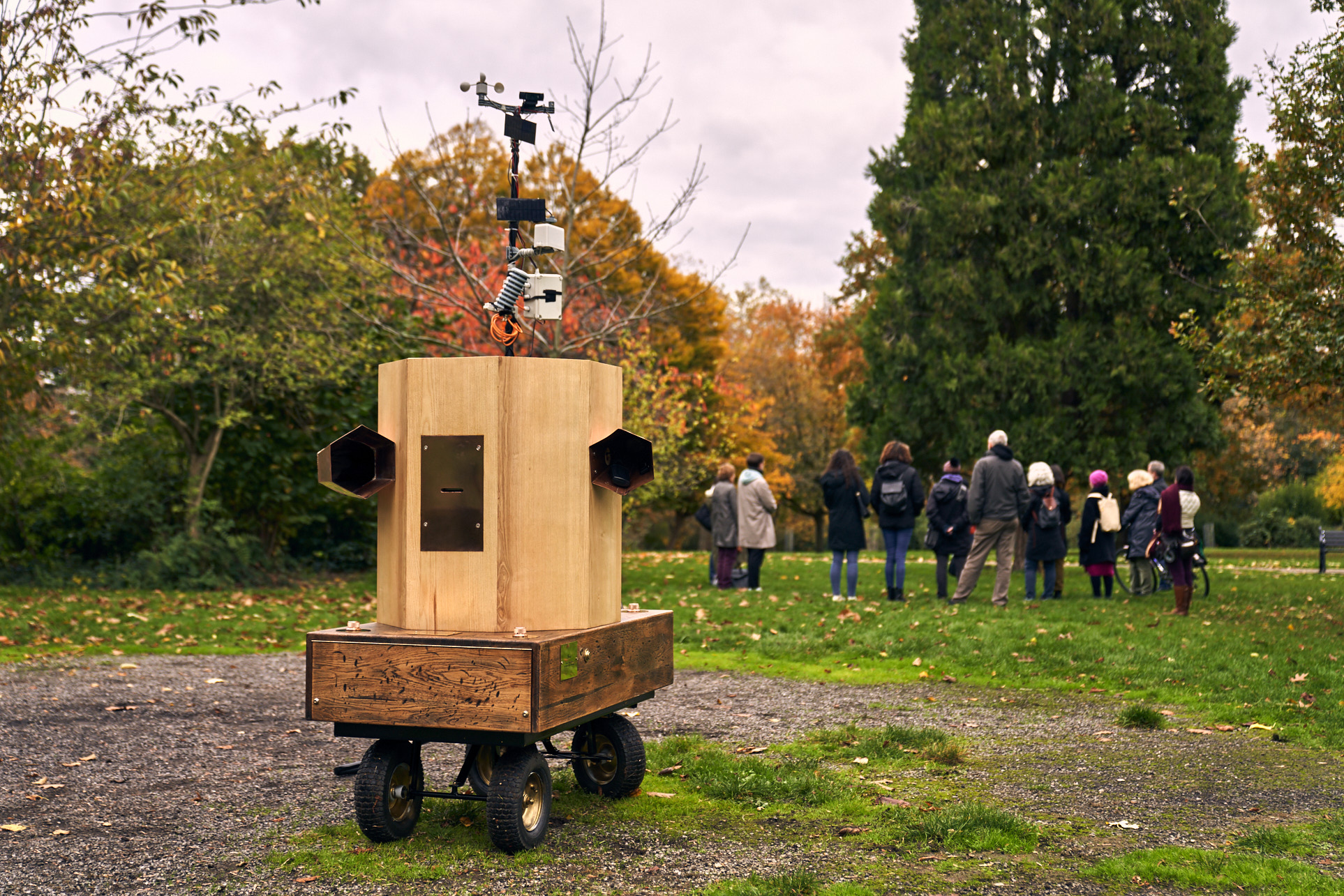 Future Machine standing in the remembrance circle in Finsbury Park, the people in the procession are in the background looking out at the Autumnal trees