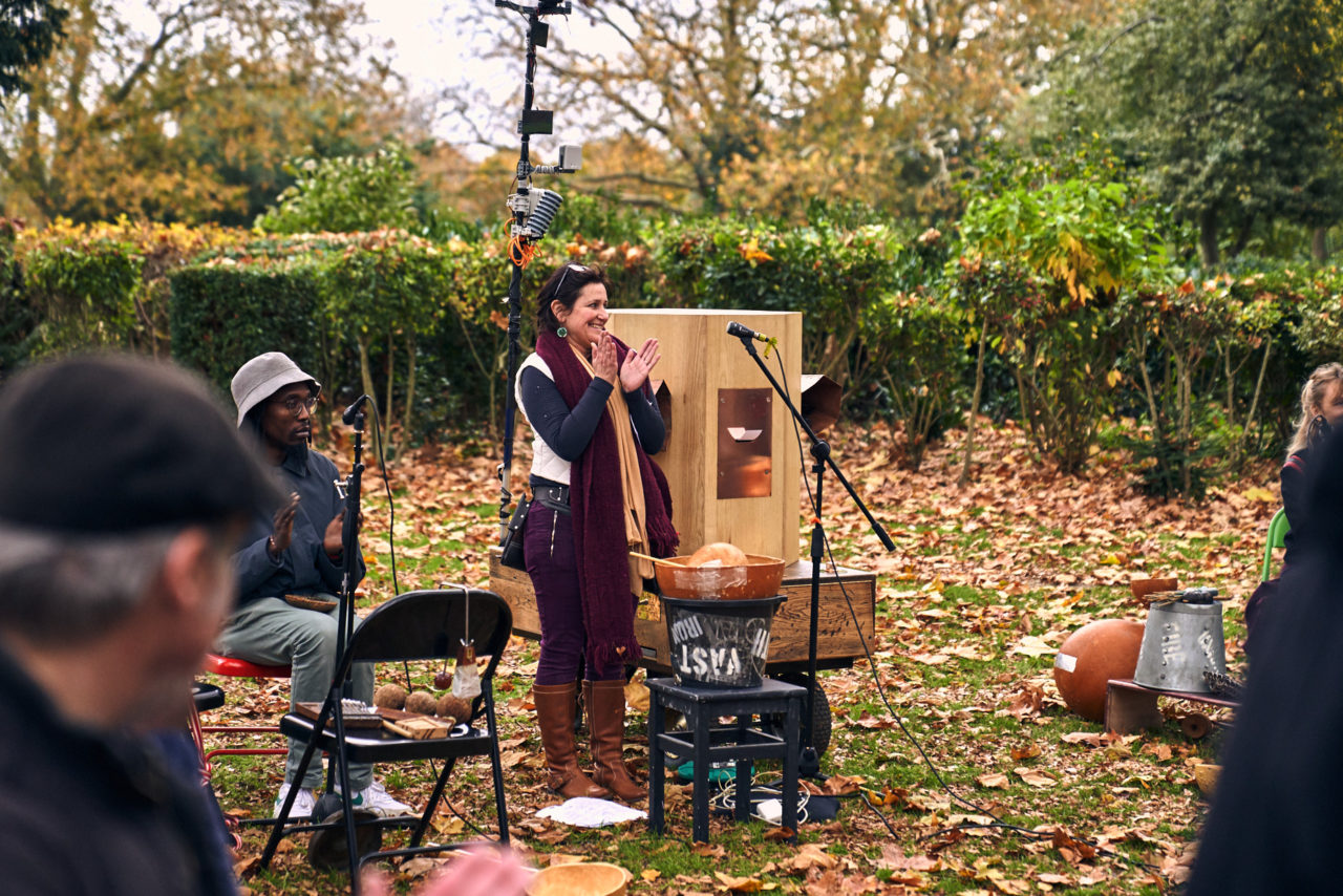 Rachel Jacobs clapping next to the Future Machine, Miles Ncube clapping surrounded by instruments and microphones and autumunal trees and leaves on the ground