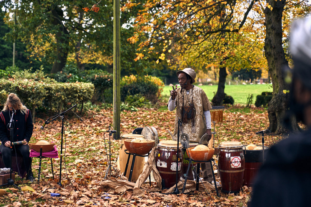 Alex speaking into a microphone next to drums, water bowls and other instruments, next to Miles Ncube surrounded by autumn leaves and trees