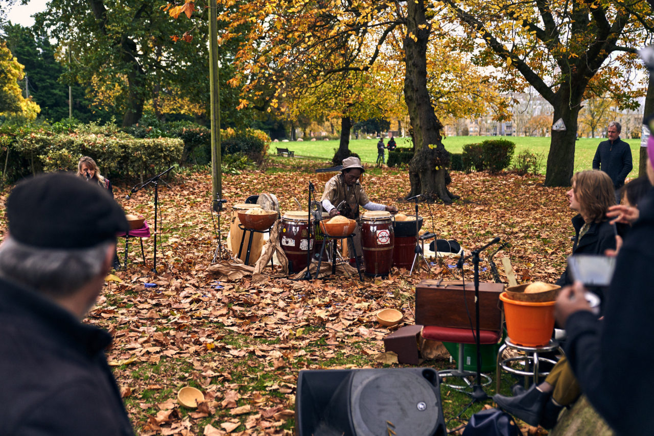 Alex drumming surrounded by autumn leaves and trees, David in front with insturments and people watching