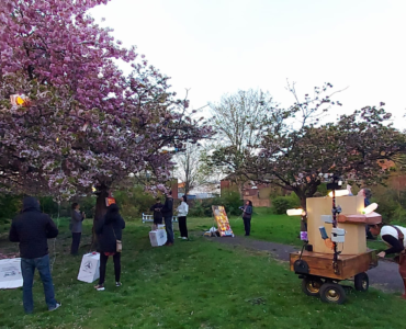 Pink blossom trees in Christ Church Gardens with light boxes in the trees, Future Machine in front with artist companion next to it and people with hand generators lighting up the trees