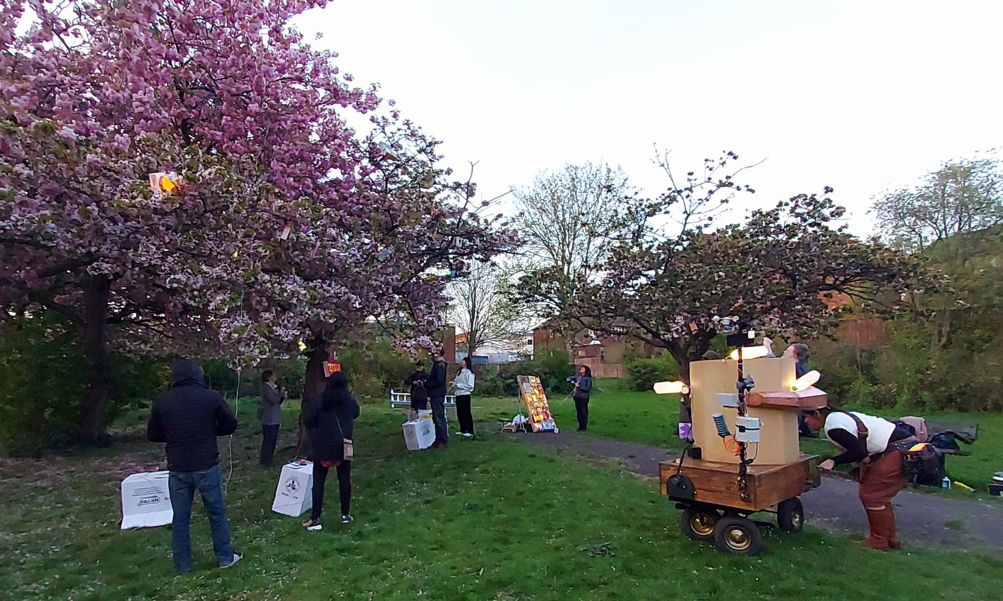 people standing around the blossom trees with lit up bird boxes in amongst the blossoms. Rachel turning the handle of the Future Machine that is also lit up with plastic white milk bottle lights