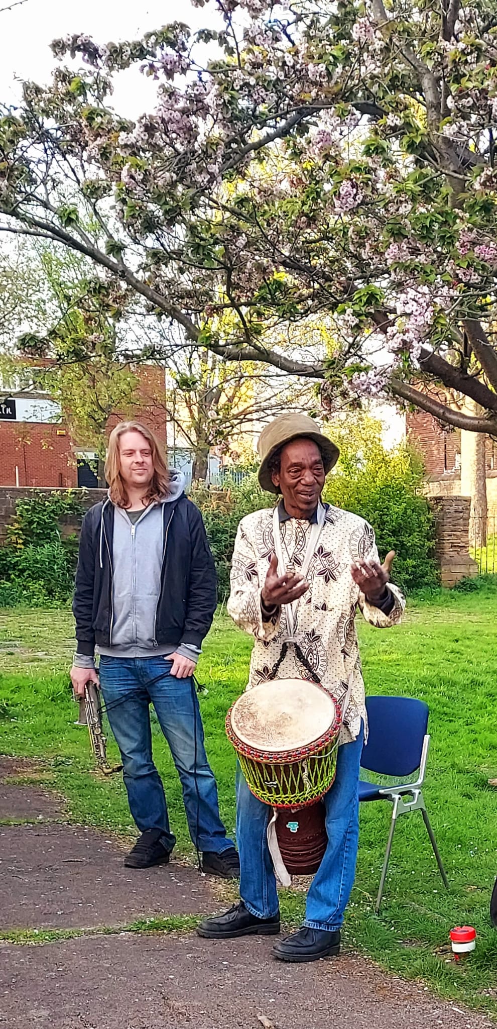 Alex with his Djeme talking and Dave behind with his saxaphone in Christ Church Gardens