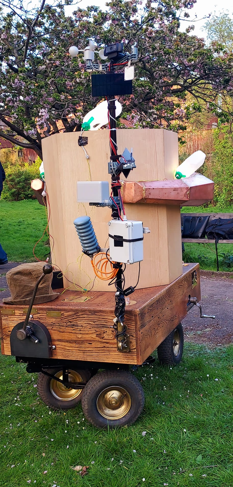 Future Machine from the back with a metal lever, a large black pole with a solar panel and weather sensors on it (wind, rain, co2, temperature), the oak base, wheels with brass middles below and the octagon on top made of ash, with the large copper trumpet to the side and the blossom tree in the distance, Plastic milk bottle lights are attached to the top and side of Future Machine and Alex's hat is sitting on the back