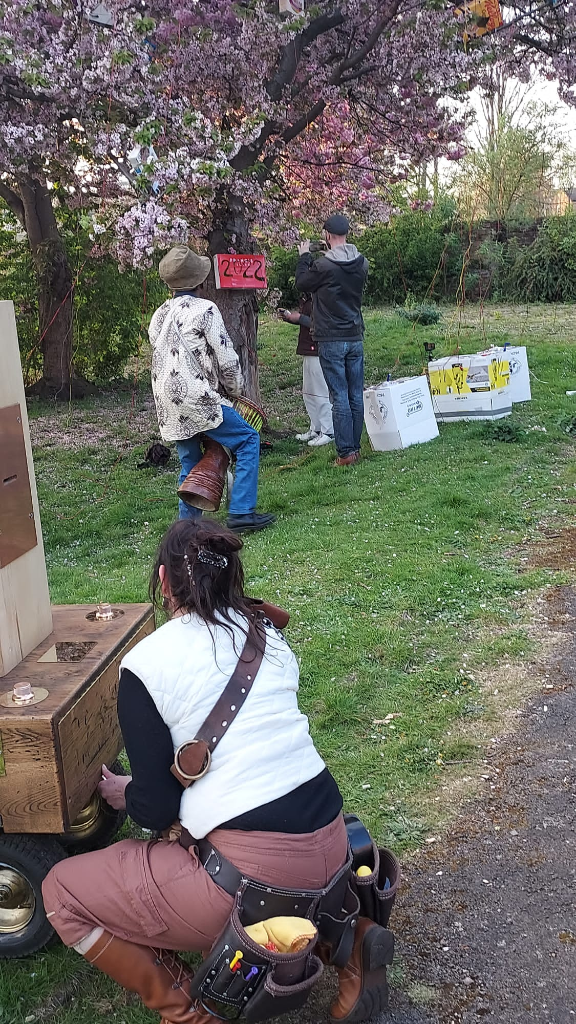 Rachel bending down next to the front of Future Machine, Alex is playing his djembe drum under the blossom tree, Robin is turning a hand generator next to the blossom tree and the red sign with 2022 cut out