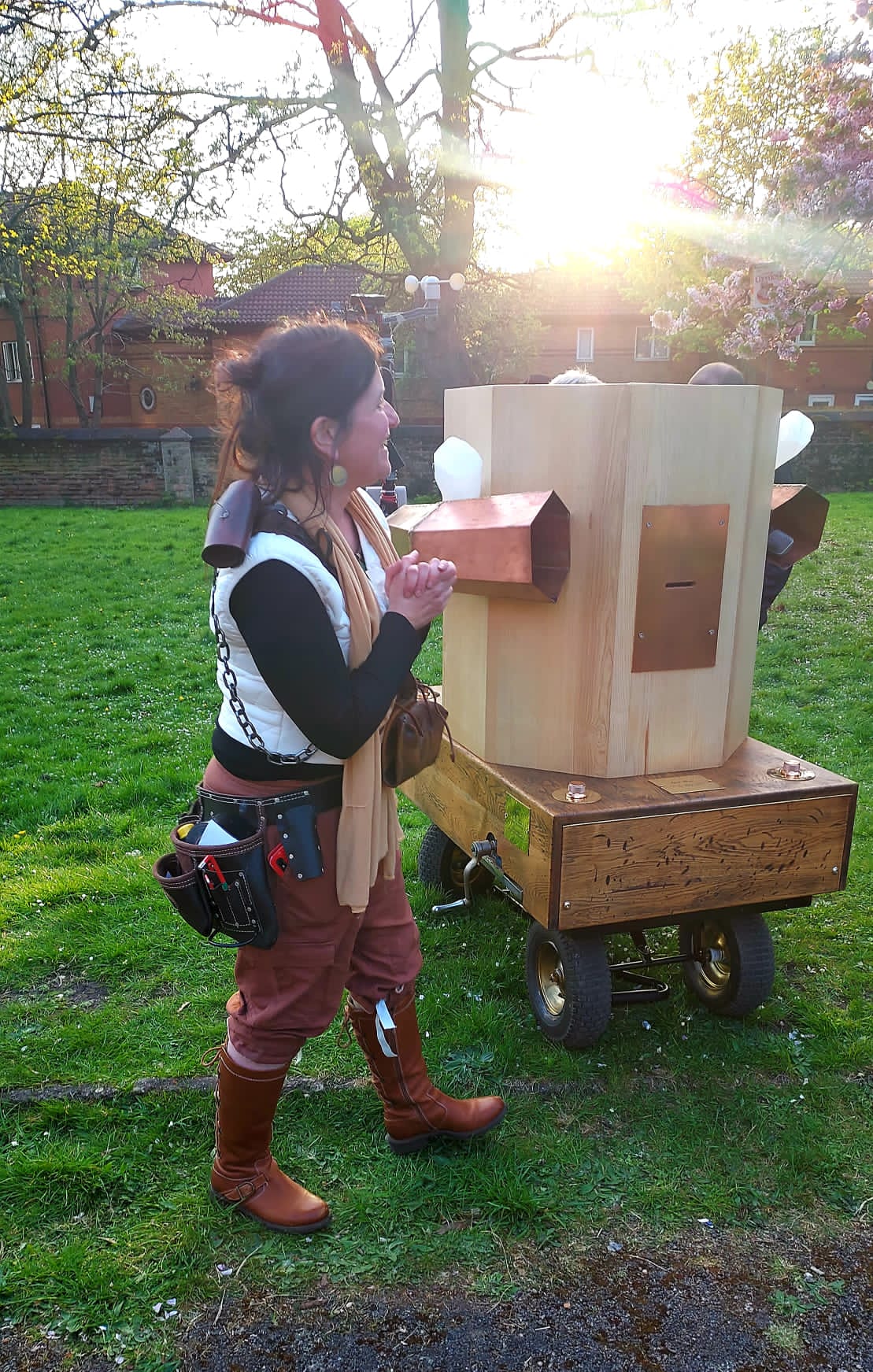 Rachel dressed in brown bloomers, brown boots, a large tool belt around her waist, a white gilet, black arms and a beige scarf. Future Machine with plastic milk bottle lights attached to both side and the top, light coming through the trees behind