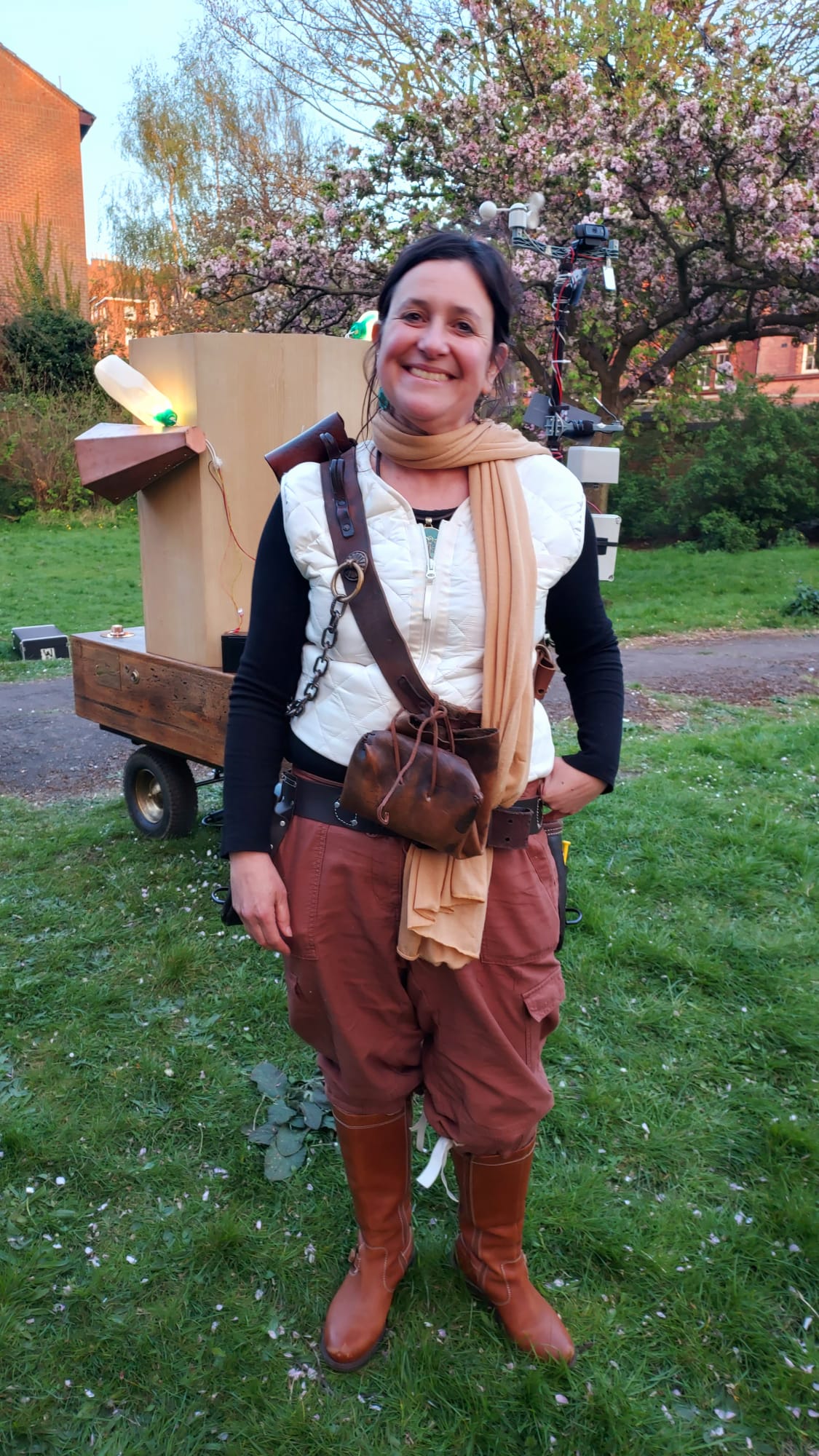 Rachel dressed in brown bloomers, brown boots, a large tool belt around her waist, a white gilet, black arms and a beige scarf. Future Machine lit up behind her and blossom trees behind the Future Machine