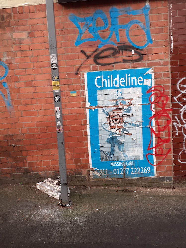 Lamp pose with stickers and a chain with a block attached to it in front of a red brick wall with a ripped poster and image of Banksy's graffiti of a girl with a tyre around her middle doing the hula and layers of graffiti tags on top