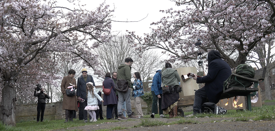 people standing around Future Machine and talking, a man playing the guitar on a bench, under the blossom trees