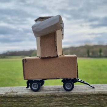 a small cardboard model of Future Machine on a fence with a field and trees and sky in the background