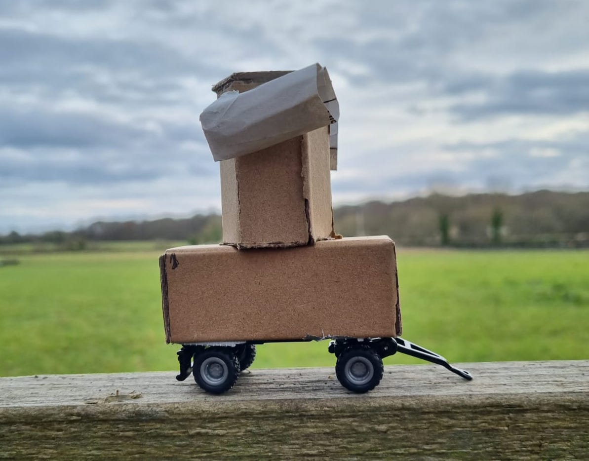 a small cardboard model of Future Machine on a fence with a field and trees and sky in the background