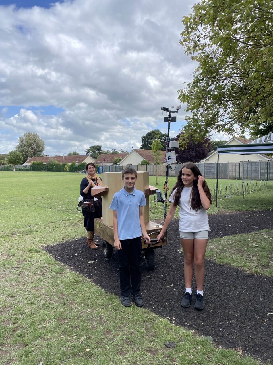 Two children pulling the Future Machine under a cloudy and blue sky with Rachel in the background