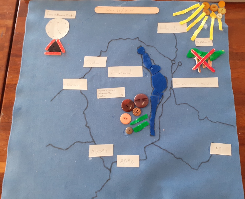 A data map made of felt with lake windermere and small signs on it naming places and roads and a sign saying time is running out with trees with a cross over them and the sun coming in at the edge