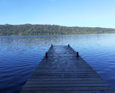 end of the wooden pier on a blue lake with a forest on a hill on the far shore and blue sky