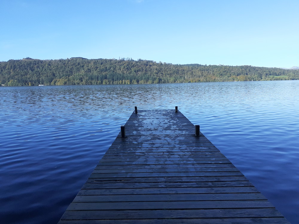 end of the wooden pier on a blue lake with a forest on a hill on the far shore and blue sky