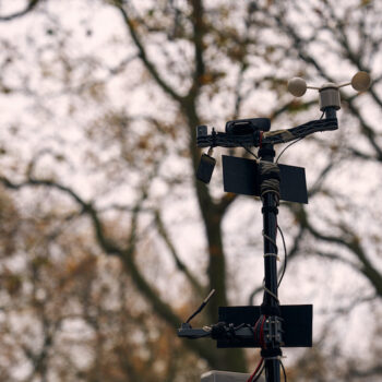 a pole with two small solar panels, a weather vane, moisture sensor and a webcam with trees in the background.