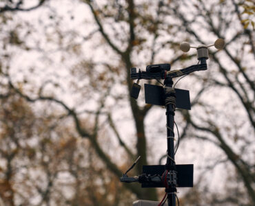 a pole with two small solar panels, a weather vane, moisture sensor and a webcam with trees in the background.